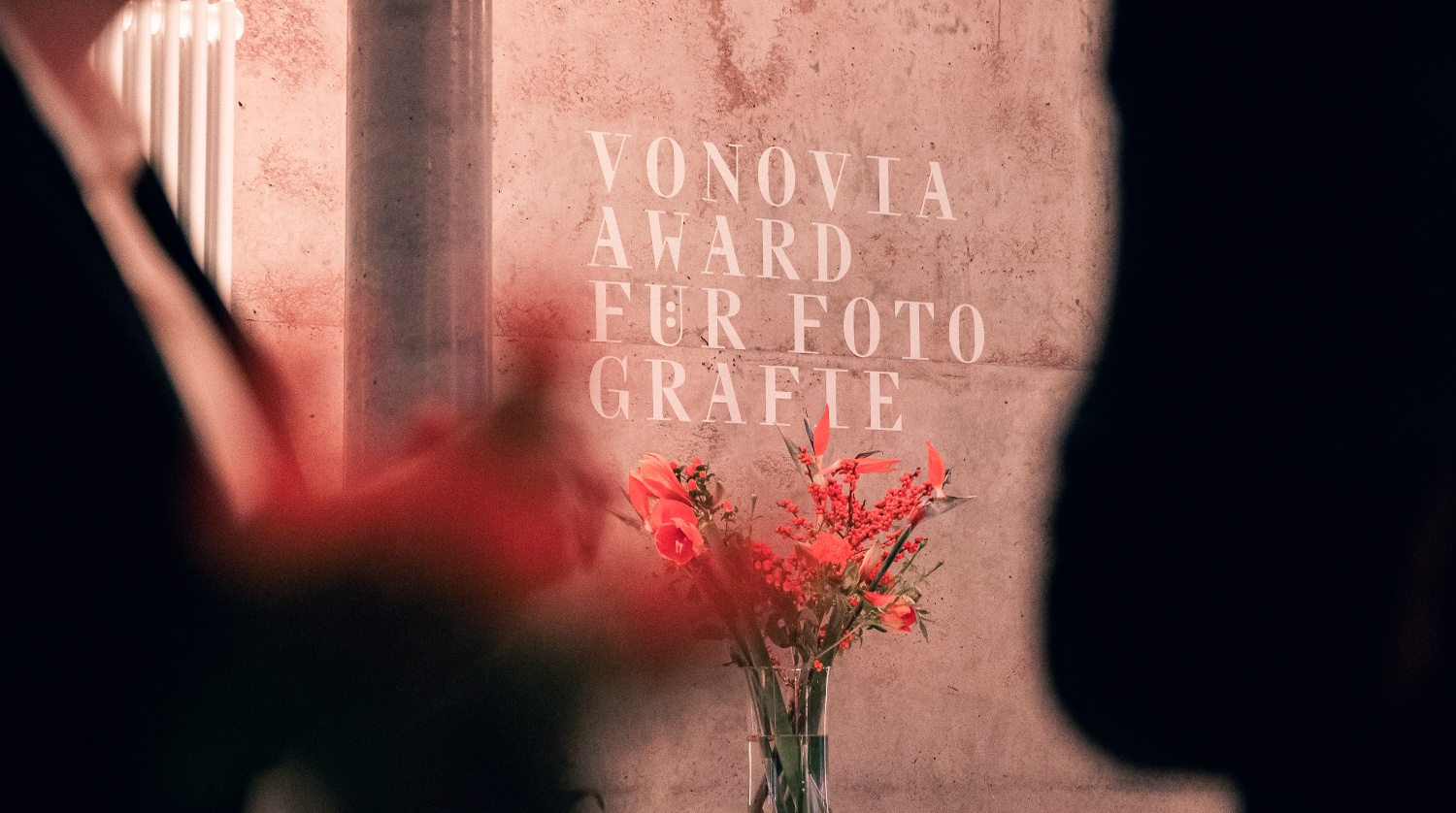 Steinwand mit Text: Vonovia Award für Fotografie. Davor eine rote Blume in einer Phase. Im Vordergrund verschwommene Menschen in dunkler Kleidung.