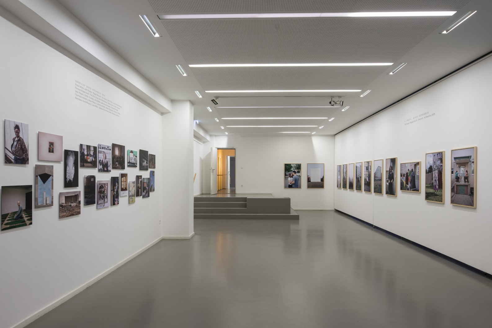 Kommunale Galerie Berlin Moderne Kunstgalerie mit weißen Wänden und grau gläzendem Boden. An beiden Seitenwänden hängen zahlreiche Fotografien. Am Ende des Raumes führt eine kleine Treppe zu einer Tür.