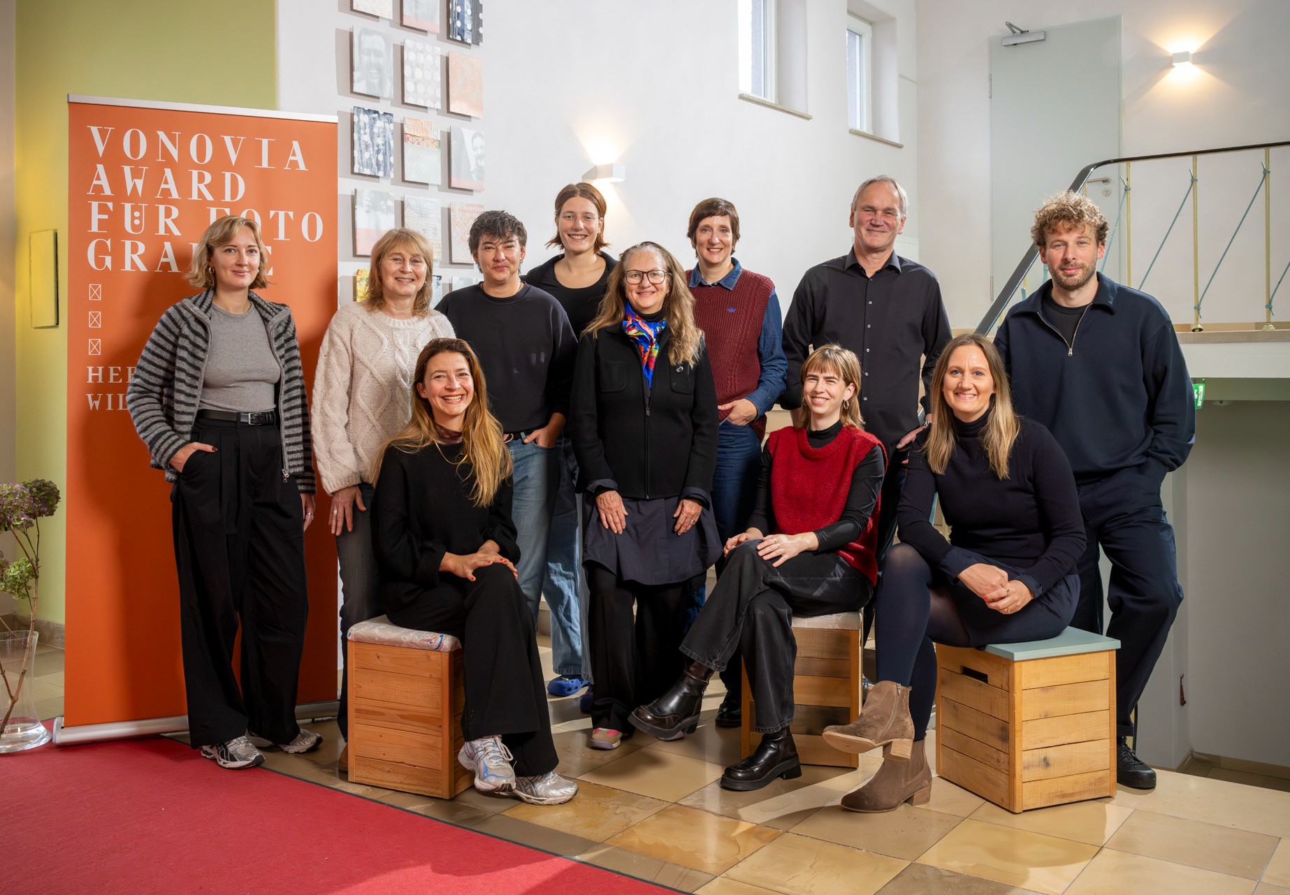 Eine Gruppe von 11 Personen steht und sitzt in einem Innenraum vor einem Banner für den Vonovia Award für Fotografie.