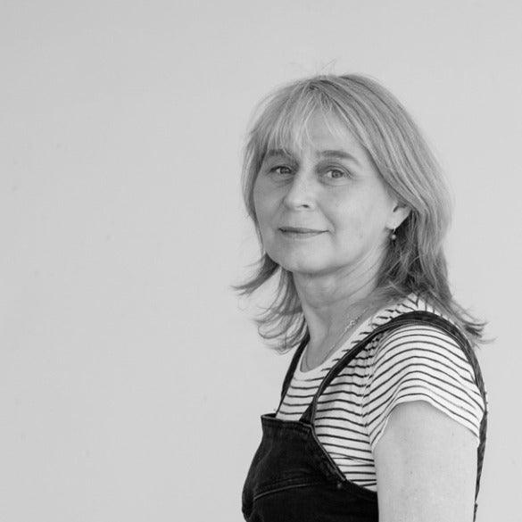 Schwarz Weiß Portrait von Ute Behrend. Frau mit mittellangem, hellem Haar und Pony. Sie trägt ein gestreiftes Oberteil und eine dunkle Latzhose, steht vor einer weißen Wand und schaut schräg in die Kamera.