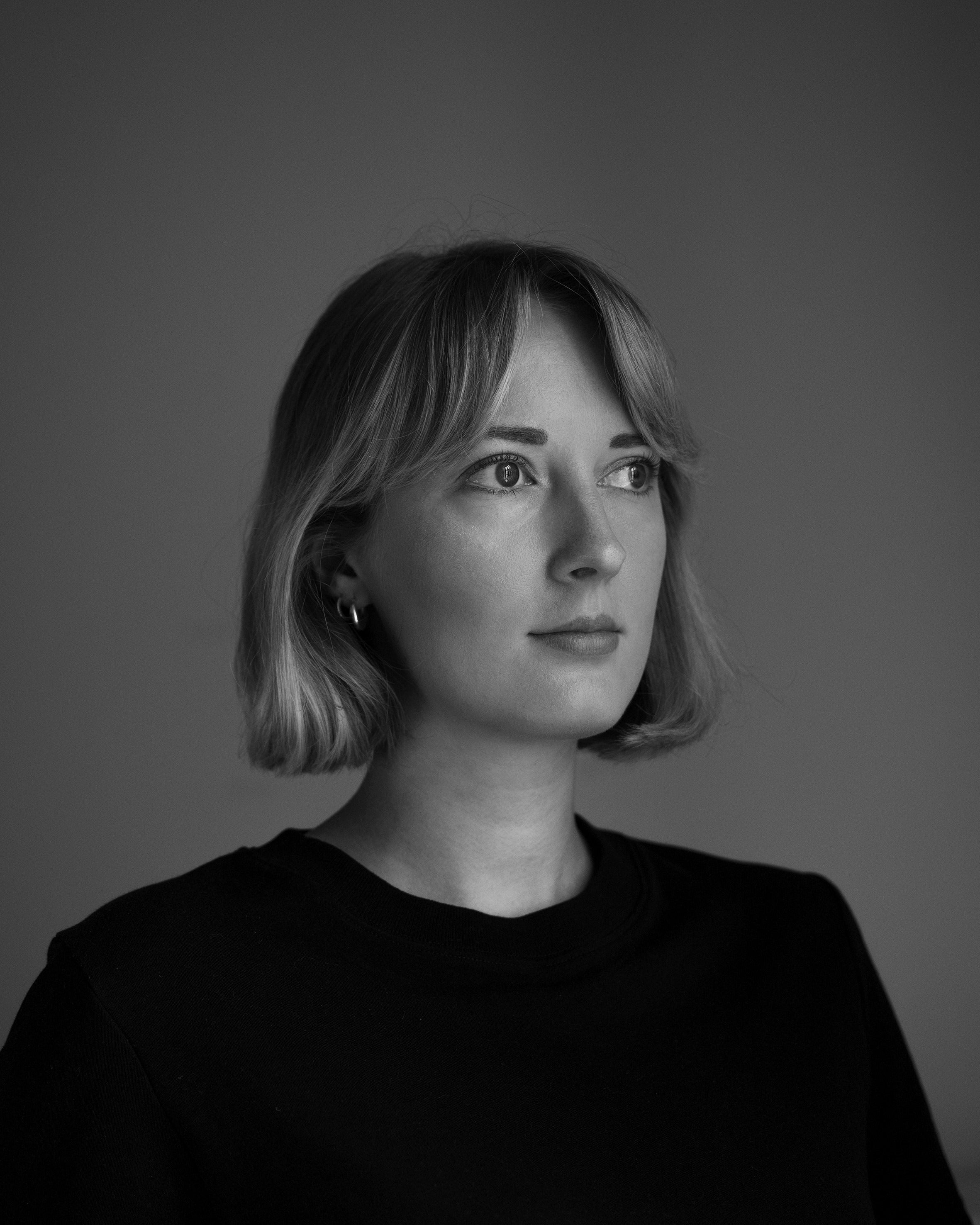 Schwarz Weiß Portrait einer jungen Frau mit einem kurzen Bob und Pony. Sie trägt ein dunkles Oberteil und blickt nach rechts.