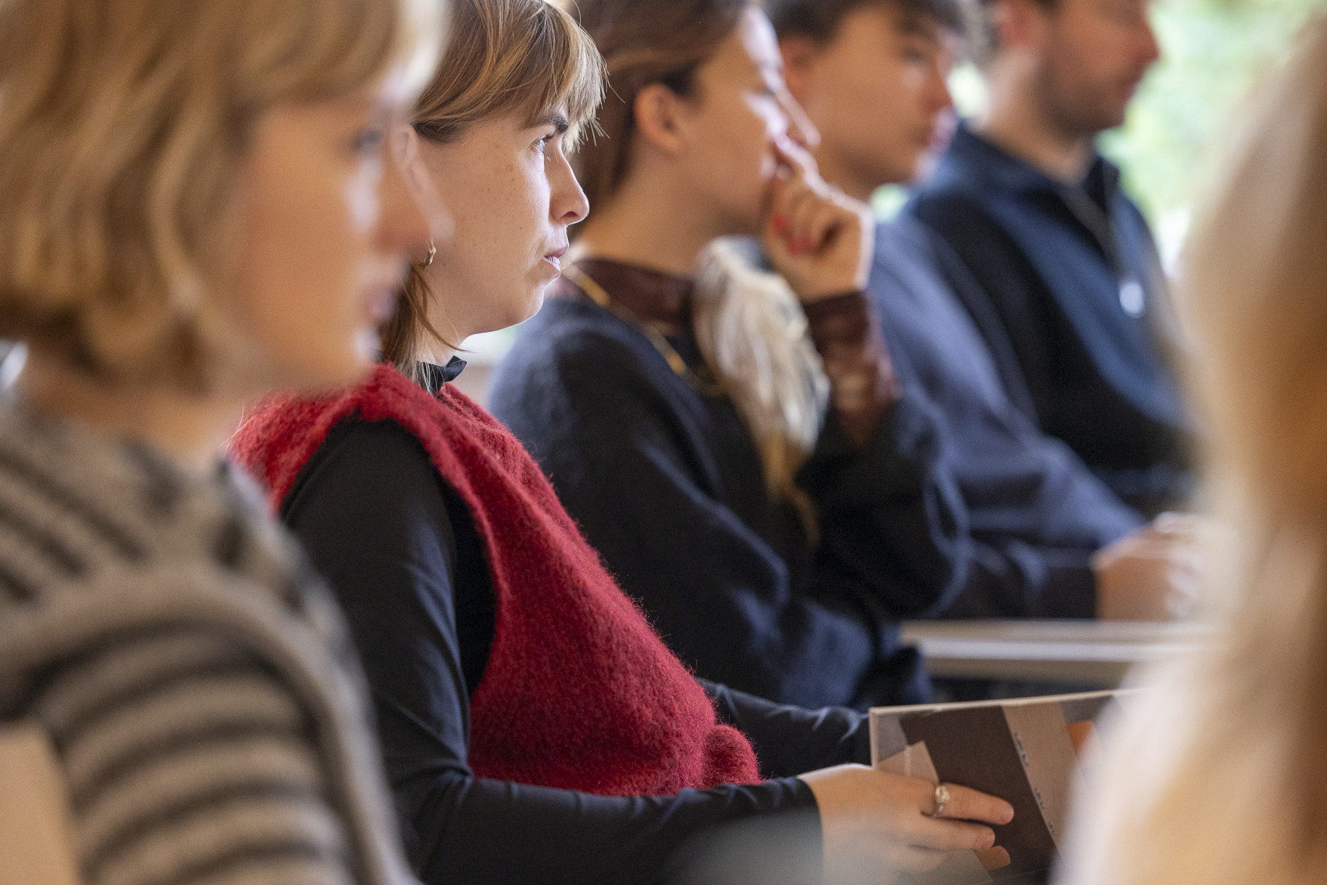 Gruppe von Personen ist im Profil sitzend zu sehen, wobei Unschärfe auf Vorder- und Hintergrund verteilt ist.