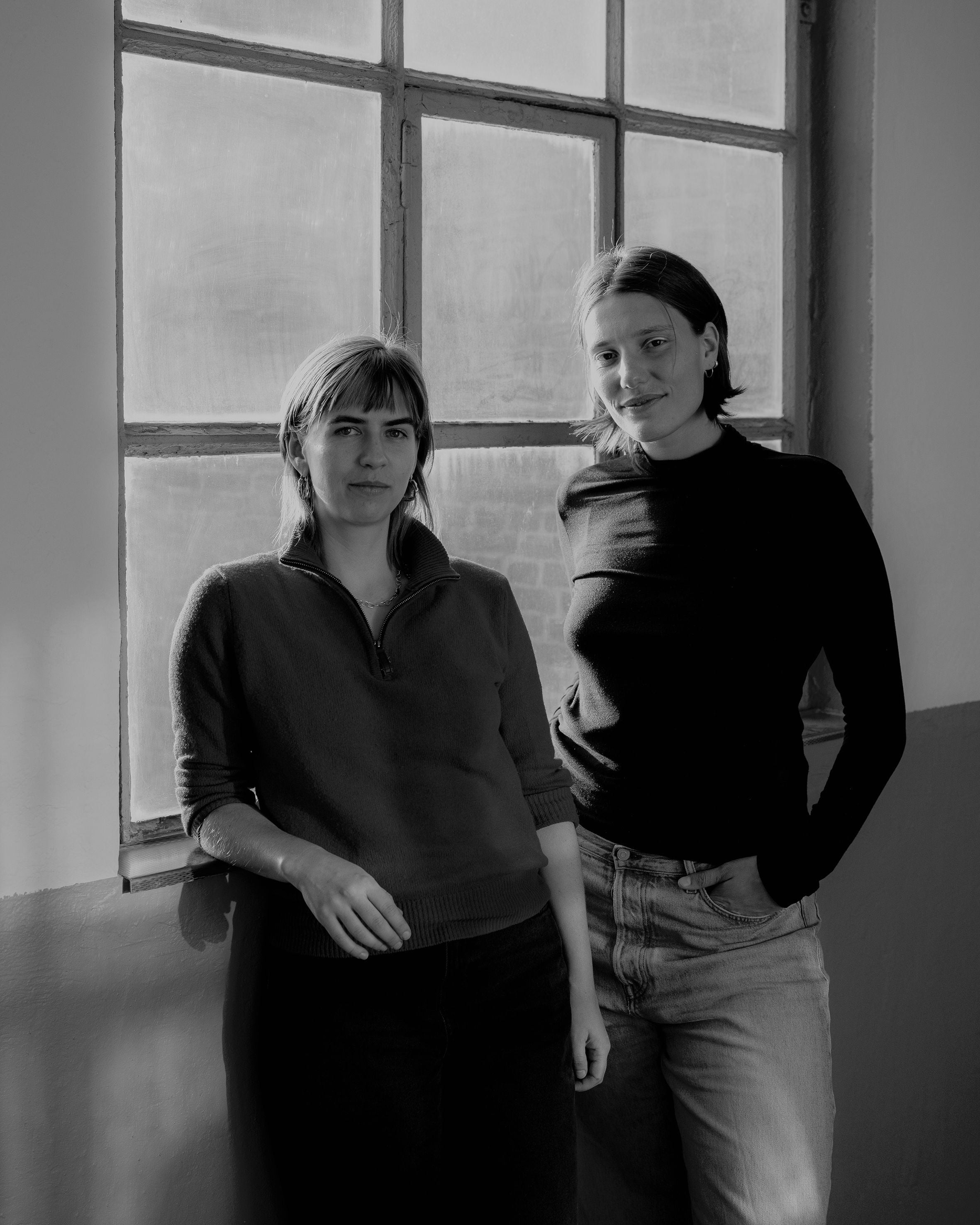 Schwarz Weiß Portrait von Stella Weiß & Rosa Burczyk. Die Beiden stehen in dunklerer Kleidung vor einem Fenster.